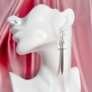 May include: Silver sword dangle earrings. The sword has a detailed design and is hanging from a silver hook.