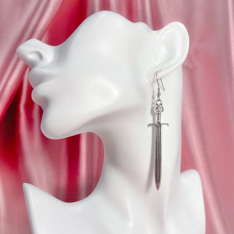 May include: Silver sword dangle earrings. The sword has a detailed design and is hanging from a silver hook.