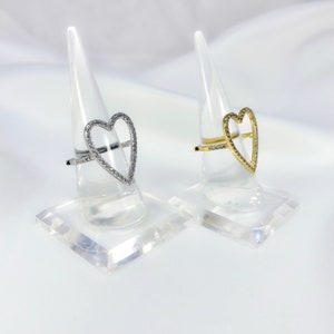 Puede incluir: Dos anillos en forma de coraz&oacute;n con un contorno de cristal transparente. El anillo de la izquierda es de plata y el anillo de la derecha es de oro.