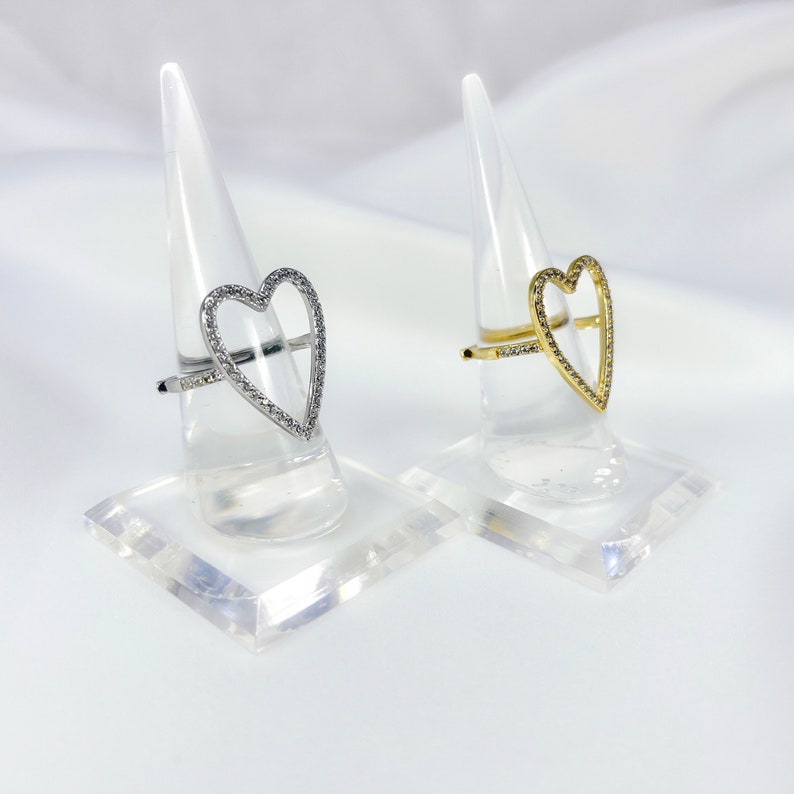 Puede incluir: Dos anillos en forma de coraz&oacute;n con un contorno de cristal transparente. El anillo de la izquierda es de plata y el anillo de la derecha es de oro.