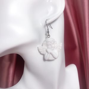 Puede incluir: Pendientes colgantes de cer&aacute;mica blanca con forma de querub&iacute;n &aacute;ngel con ganchos plateados. El &aacute;ngel tiene las manos juntas y mira hacia abajo.