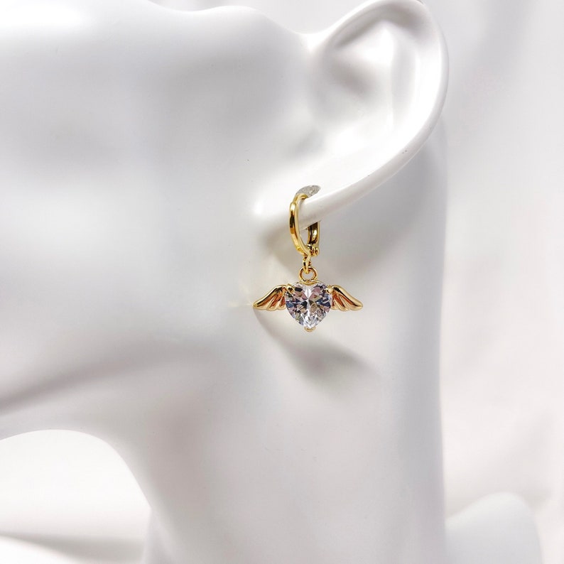 May include: Gold hoop earrings with a dangling heart-shaped crystal accented with gold wings.