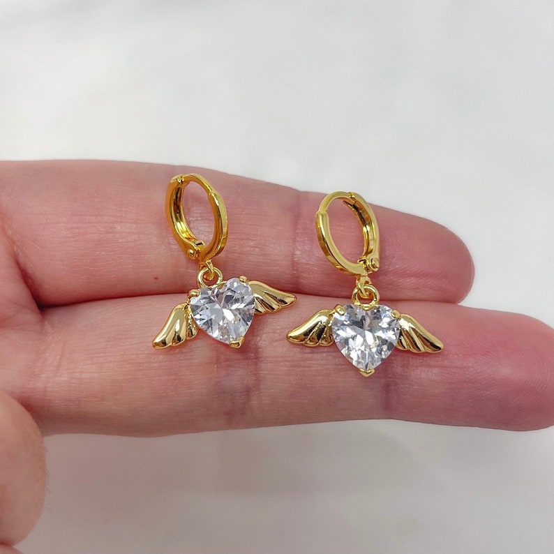 May include: Gold hoop earrings with a dangling heart-shaped crystal charm with small wings on each side.
