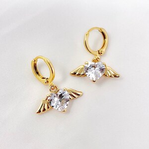 May include: A pair of gold hoop earrings with a dangling heart-shaped crystal and angel wings.