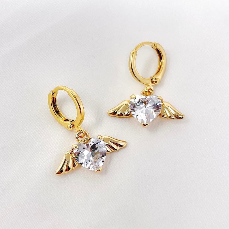 May include: A pair of gold hoop earrings with a dangling heart-shaped crystal and angel wings.