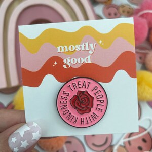 K&ouml;nnte beinhalten: Eine rosa und wei&szlig;e Emaille-Anstecknadel mit einer roten Rose und dem Text "Treat People With Kindness". Die Anstecknadel befindet sich auf einer Karte mit dem Text "mostly good" in einem welligen Design in Rosa und Gelb.
