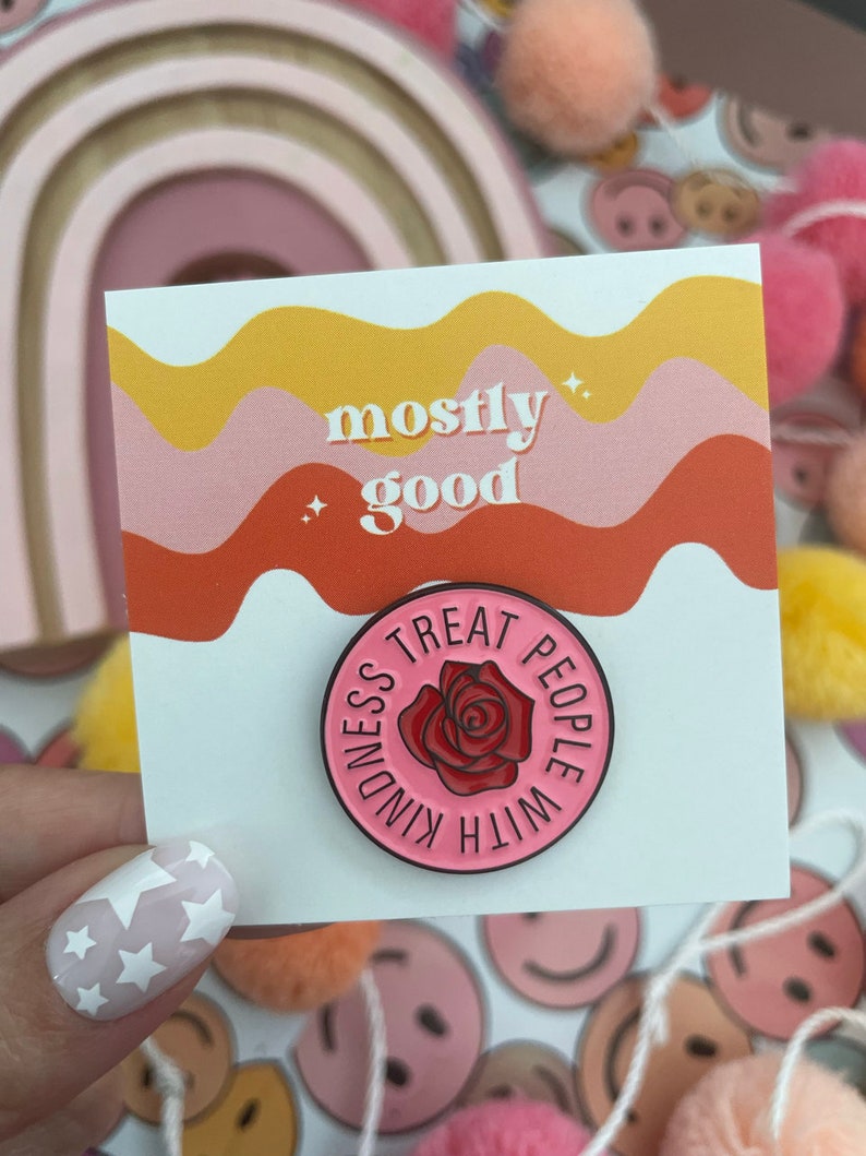 K&ouml;nnte beinhalten: Eine rosa und wei&szlig;e Emaille-Anstecknadel mit einer roten Rose und dem Text "Treat People With Kindness". Die Anstecknadel befindet sich auf einer Karte mit dem Text "mostly good" in einem welligen Design in Rosa und Gelb.