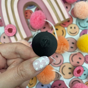 K&ouml;nnte beinhalten: Schwarze Emaille-Anstecknadel mit silbernem Metallverschluss. Die Anstecknadel zeigt ein wei&szlig;es Sternendesign. Die Anstecknadel wird vor einem Hintergrund aus bunten Bommeln und Smiley-Gesichtern gezeigt. Der Text auf der Anstecknadel lautet "Treat People With Kindness".