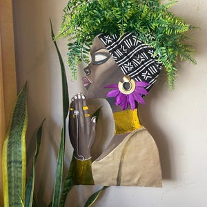 May include: A wooden wall art piece depicting a woman's profile with a black and white head wrap, gold earrings, and a purple flower. The woman's neck is adorned with gold, and her hands are clasped in prayer, wearing rings and a bracelet. Green foliage forms a crown.