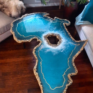 May include: A teal blue resin coffee table shaped like the continent of Africa. The table has a gold glitter edge and a gold glitter line down the center. The table has a large hole in the center, which is filled with white resin and gold glitter.