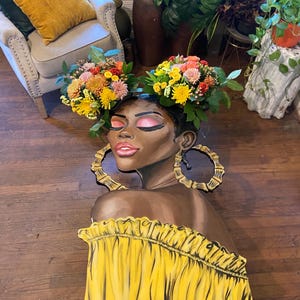May include: A decorative table featuring a painted portrait of a woman with floral headpieces and large gold hoop earrings. The woman is wearing a yellow off-the-shoulder top. The table has black metal legs and is placed on a wooden floor.