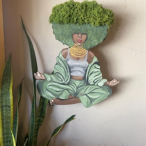 May include: A wooden wall art piece depicting a person in a meditative pose. The figure is painted in shades of green, wearing a gold necklace and a white top. The hair is made of green moss. The art piece is mounted on a beige wall.