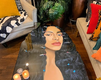 Peut inclure: Une table basse noire avec un plateau en résine représentant le portrait d'une femme aux longs cheveux noirs. La table a un cadre en métal noir et est décorée de petites touches bleues.