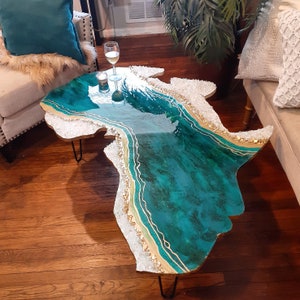 May include: A teal and white resin coffee table with gold accents and a unique, organic shape. The table has a glossy finish and is supported by hairpin legs.