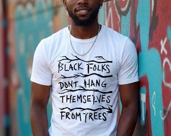 May include: A white short-sleeved t-shirt featuring black text that reads "BLACK FOLKS DONT HANG THEMSELVES FROM TREES" with a graphic of tree branches integrated into the lines of text, worn with a silver chain necklace and blue jeans.