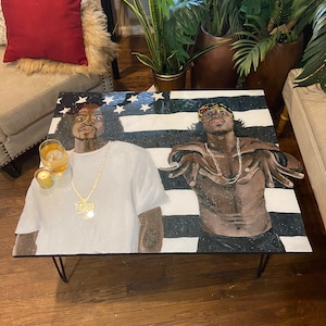 May include: A square coffee table with a painted design featuring two men in front of a black and white striped background with stars. The table has black metal hairpin legs. A glass of liquid and a candle are on the table.