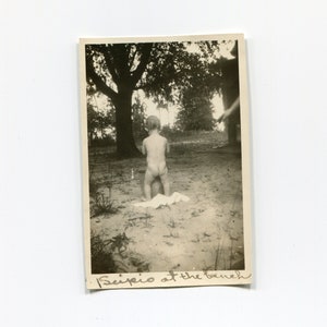 May include: A vintage black and white photograph of a young child standing on a sandy surface near a tree. The child is facing away from the camera. The handwritten text at the bottom reads "Scipio at the beach."