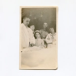 May include: Black and white photo of a group of people, including a doctor, a nurse, and three children. The doctor is wearing a white coat and is looking at the camera. The nurse is standing behind the doctor and is looking at the camera. The children are standing in front of the doctor and are looking at the camera. The photo is taken in a hospital room.
