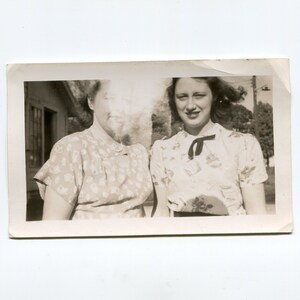 May include: Two women in vintage clothing stand side-by-side, smiling at the camera. The woman on the left is wearing a floral print dress with a white collar. The woman on the right is wearing a white blouse with a black bow tie and a floral print.
