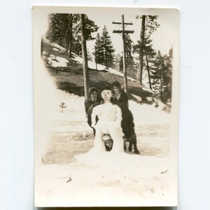 May include: A black and white photo of four people building a snowman in a snowy forest. The snowman is wearing a hat and has a face.