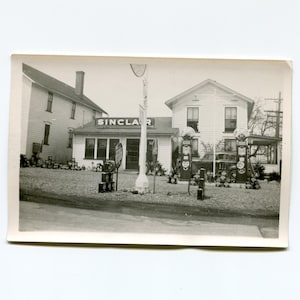 May include: A black and white photo of a Sinclair gas station. The station has a sign that says "Sinclair" and two gas pumps. There is a car parked in front of the station.