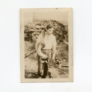 Peut inclure: Une photographie en noir et blanc d'un homme tenant un grand morceau de bois courbé. Il porte un t-shirt blanc et un pantalon foncé. L'arrière-plan est un décor rocheux en plein air.