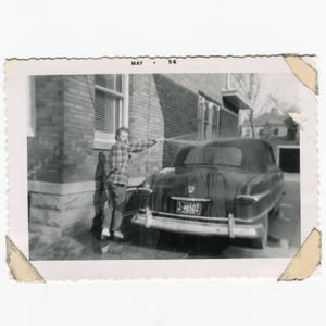 May include: Vintage black and white photograph of a person washing a classic car. The car has a license plate and is parked near a brick building. The photo is dated May '56.