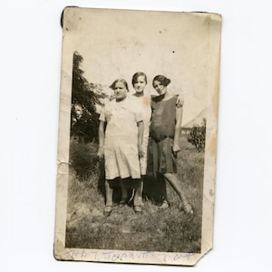 May include: Black and white photo of three women standing in a grassy field. The women are wearing dresses and are smiling. The photo is dated "July 7, 1927".