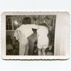 May include: A vintage black and white photograph with a scalloped edge showing three individuals standing together with their arms around each other. The central figure wears a light-colored shirt and dark trousers, while the others wear light tops.