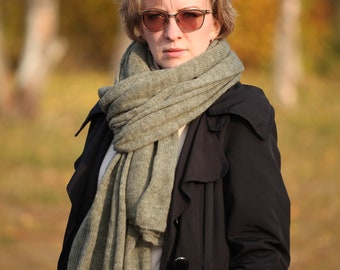 May include: A woman wearing a black coat and a gray scarf, with sunglasses on her face, standing in a field with a blurred background of trees.