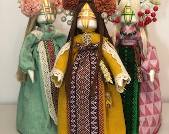 May include: Three handmade fabric dolls wearing traditional clothing and floral headbands. The dolls are standing on a white surface. The doll in the center is wearing a yellow dress with a brown and red patterned sash. The doll on the left is wearing a green dress with a white lace trim. The doll on the right is wearing a pink and green dress with a white lace trim.