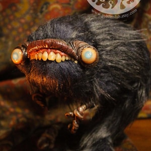 May include: A furry, dark grey creature with large, blue eyes and a toothy grin. The creature has a long, bushy tail and is sitting on a patterned rug. The image includes the text "Goblins Lab" and a website address.