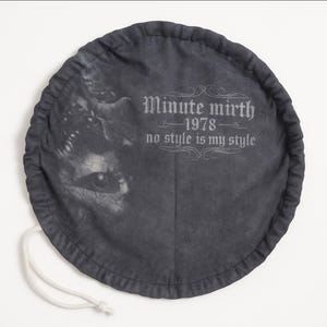May include: A circular dark grey fabric pouch with a drawstring closure. The surface features a faded graphic of a face on the left and the printed text "Minute mirth 1978 no style is my style" in a decorative font on the right side.