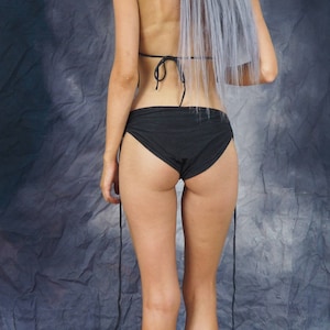 Puede incluir: Una mujer con un bikini negro de tiro bajo con tirantes finos. El bikini est&aacute; atado a los lados. La mujer tiene el pelo largo, liso y plateado.