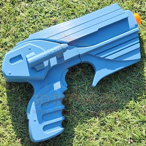 Peut inclure: Un pistolet jouet bleu clair avec un embout orange, posé sur de l'herbe verte. Le pistolet a un design futuriste avec une poignée, une gâchette et un canon. Le corps du pistolet présente une série de lignes parallèles et une petite fixation cylindrique.