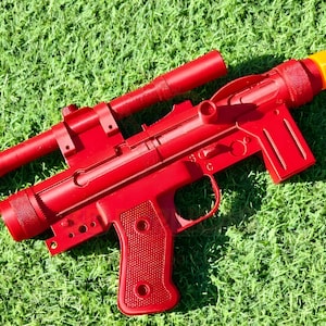 May include: A bright red toy blaster gun with a scope and an orange tip, resting on a bed of green grass. The toy gun has a detailed design, including a grip, trigger, and barrel.