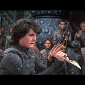 May include: A man with dark hair and blue eyes holds a silver dagger in his right hand. He is wearing a dark gray tunic and is surrounded by other people in similar attire.