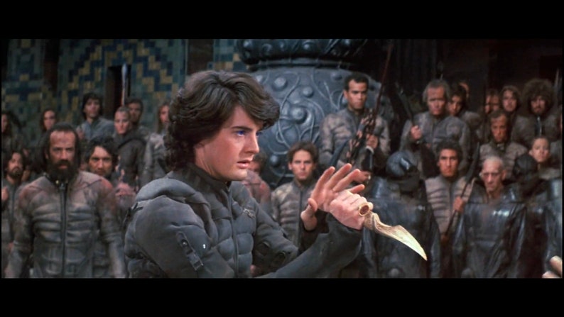 May include: A man with dark hair and blue eyes holds a silver dagger in his right hand. He is wearing a dark gray tunic and is surrounded by other people in similar attire.
