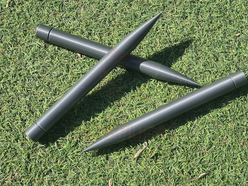 May include: Three gray metal stakes with pointed ends. The stakes are lying on a bed of green grass.