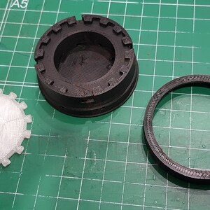 May include: Three 3D printed parts, one white and two black, with intricate designs. The white part is a circular disc with a textured surface. The black parts are a ring and a cylindrical base with a raised rim.
