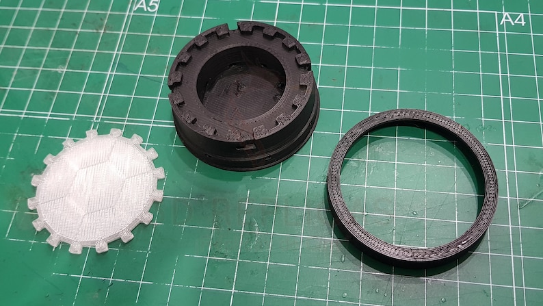 May include: Three 3D printed parts, one white and two black, with intricate designs. The white part is a circular disc with a textured surface. The black parts are a ring and a cylindrical base with a raised rim.
