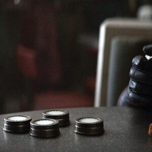 May include: Four small, round, silver-colored containers with white tops are arranged in a row on a gray table. A person's gloved hand is visible in the background.
