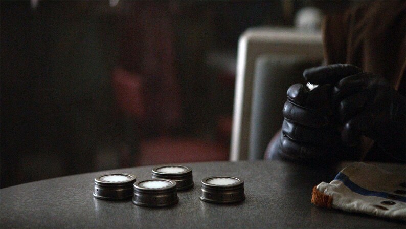 May include: Four small, round, silver-colored containers with white tops are arranged in a row on a gray table. A person's gloved hand is visible in the background.
