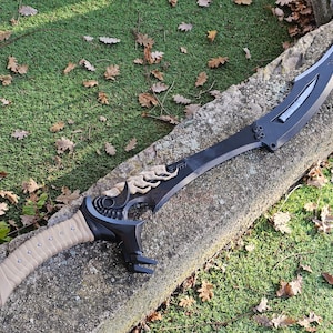May include: A black and tan fantasy sword with a curved blade and ornate handle. The sword features a tan grip, decorative bone-coloured accents, and a red button near the pommel. The blade has a silver accent and is set against a stone surface.
