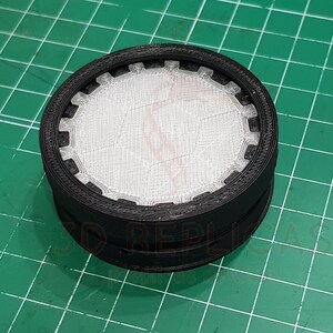 May include: A black plastic filter with a white mesh insert. The filter has a circular shape and a textured outer edge.