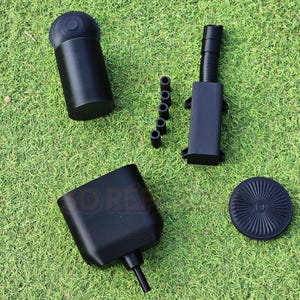 Könnte beinhalten: Eine Ansammlung schwarzer Kunststoffkomponenten, darunter ein zylindrisches Teil, ein quadratisches Teil mit einer Düse, ein geripptes Rohr, ein kreisförmiges Teil und mehrere kleine zylindrische Teile, sind auf einer grünen, grasbewachsenen Oberfläche angeordnet.