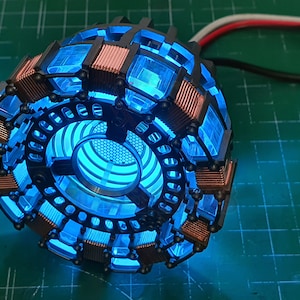 May include: A close-up of a blue and copper-coloured Iron Man Arc Reactor replica. The reactor is lit up with a bright blue light.