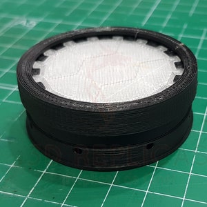 May include: A black 3D printed round object with a white mesh insert. The object has a series of small holes around the bottom edge.
