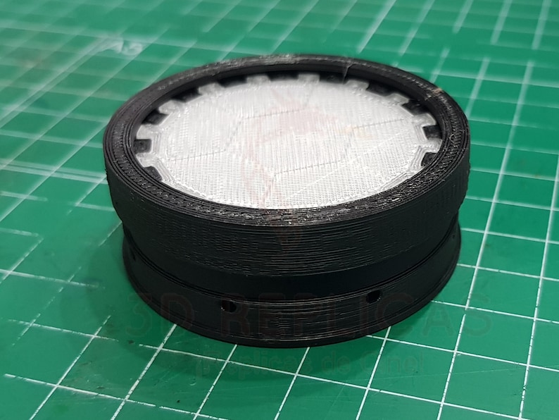 May include: A black 3D printed round object with a white mesh insert. The object has a series of small holes around the bottom edge.