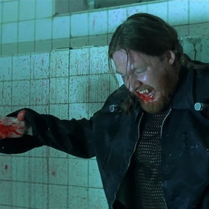 May include: A man with long hair and a beard is covered in blood. He is wearing a black leather jacket and a chainmail shirt. He is standing in a tiled room with blood splattered on the walls.